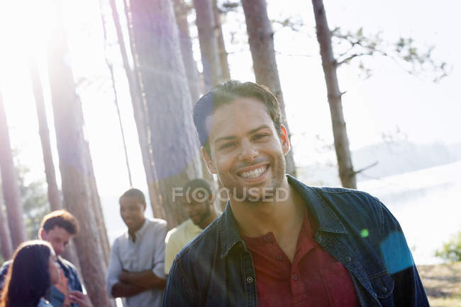 Young man smiling and looking in camera with friends gathered in pine trees at lakeside. — Stock Photo