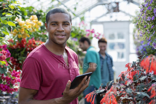 Man with digital tablet standing in greenhouse of plant nursery with coworkers in background. — Stock Photo