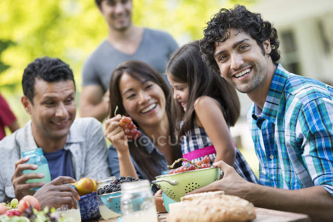 Adultos con niña de edad elemental sentados a la mesa con frutas en el jardín . - foto de stock