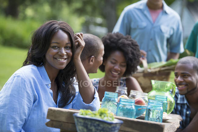 Donna sorridente e guardando in macchina fotografica con gli amici al tavolo da picnic nel giardino di campagna . — Foto stock