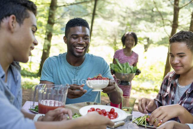 Young man holding bowl of fresh strawberries at picnic table with family in woods. — Stock Photo