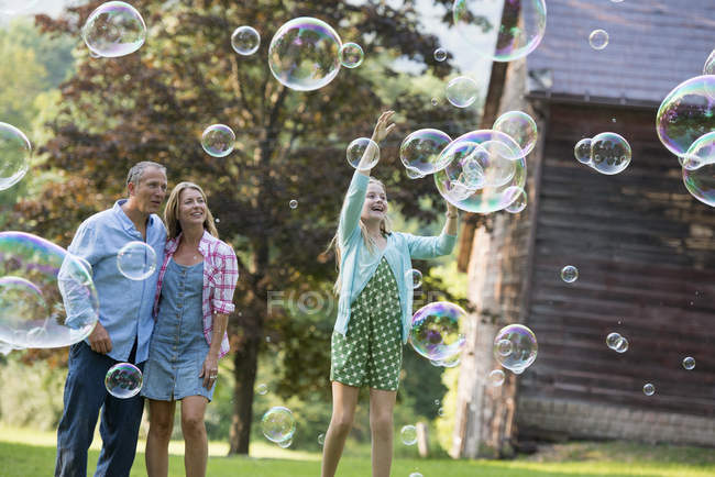 Padres viendo a su hija atrapando burbujas de jabón y riendo en el campo . - foto de stock