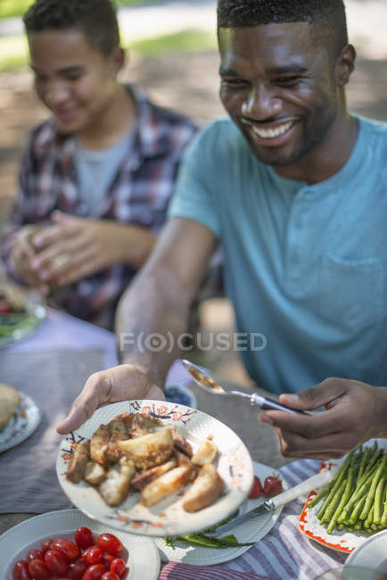 Young man holding plate of fried vegetables at picnic table with family in woods. — Stock Photo