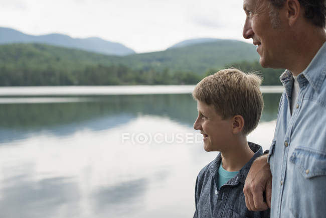 Padre e hijo de pie al aire libre en la orilla del lago y mirando a la vista . - foto de stock