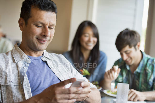Mid adult man using smartphone at cafe table with people eating in background. — Stock Photo