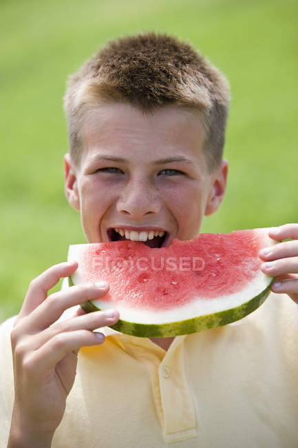 Adolescent garçon prendre morsure hors de pastèque tranche à l'extérieur et regarder dans caméra . — Photo de stock