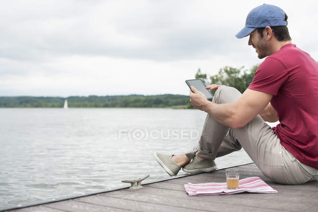 Side view of man sitting on jetty by lake and using digital tablet. — Stock Photo