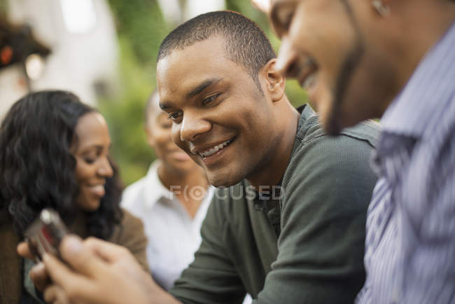 Lächelnde Männer, die mit Frauen im Hintergrund auf das Smartphone schauen. — Stockfoto