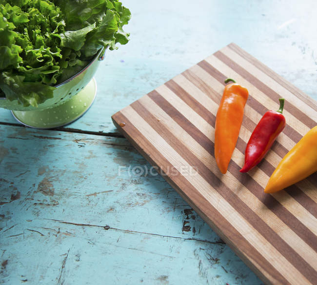 Tabletop with wooden chopping board, sweet peppers and bowl of salad leaves. — Stock Photo
