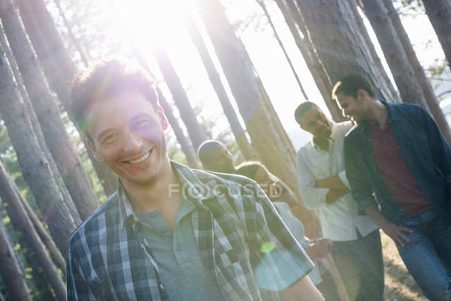 Mittlerer erwachsener Mann lächelt und blickt in die Kamera mit Freunden, die in Kiefern am Seeufer versammelt sind. — Stockfoto