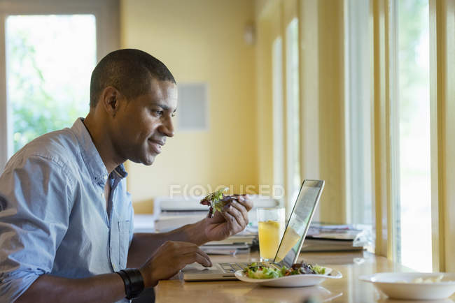 Hombre trabajando en el ordenador portátil mientras come en la mesa de la cafetería
. - foto de stock
