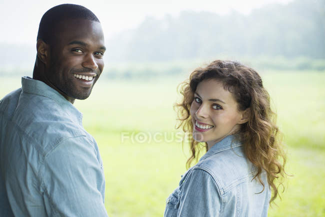 Молодая пара стоит бок о бок в зеленом поле и смотрит через плечи . — стоковое фото