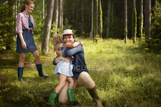 Man hugging elementary age daughter with woman watching in woodland. — Stock Photo