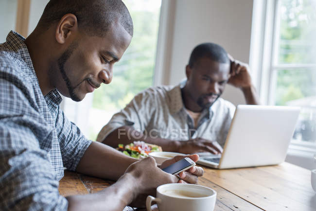 Dos hombres usando portátil y teléfono inteligente en la mesa en la cafetería . - foto de stock