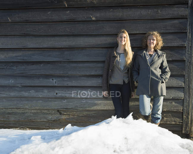 Junge und reife Frauen lehnen an Holzscheune und schauen im Winter in die Kamera. — Stockfoto