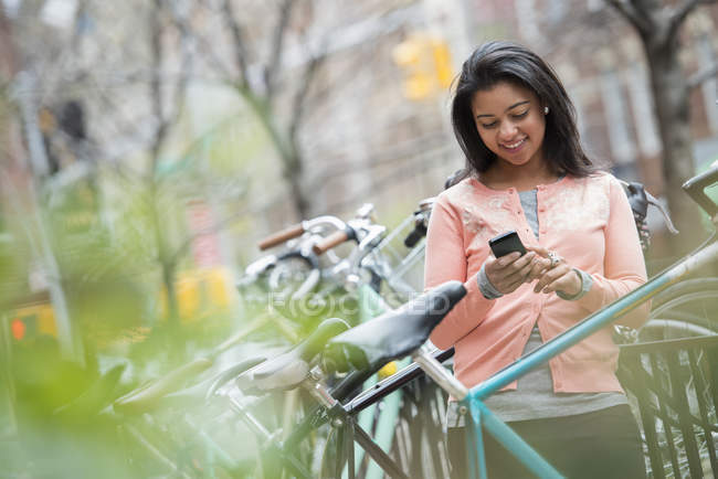 Mujer joven usando teléfono inteligente por bicicletas estacionadas en rack de bicicletas . - foto de stock