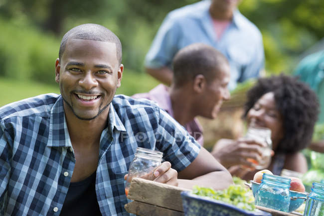 Hombre con vidrio sonriendo en la cámara con amigos en la mesa de picnic en el jardín del campo
. - foto de stock