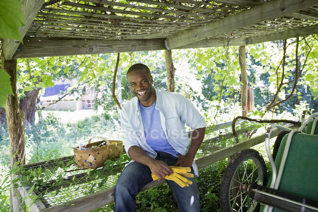 Young man sitting on bench by wheelbarrow on organic farm. — Stock Photo