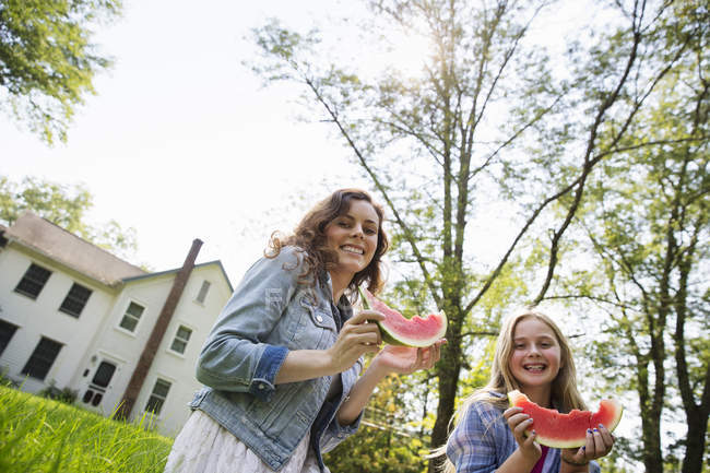 Low angle view of young woman and girl eating watermelon in farmhouse green garden. — Stock Photo