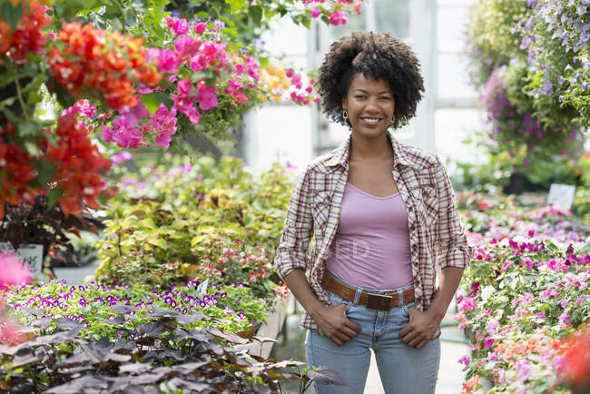 Mujer parada en vivero de plantas rodeada de plantas con flores y follaje verde . - foto de stock