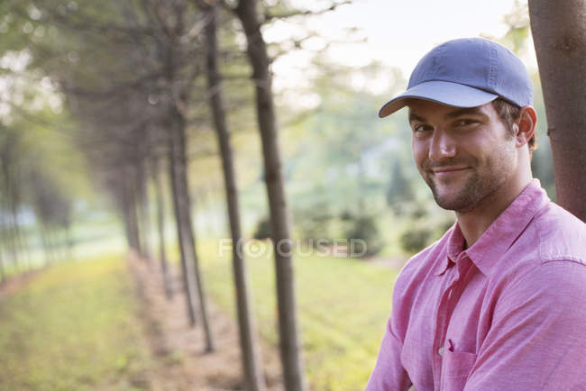 Hombre con gorra apoyado en el árbol en el parque y mirando en la cámara . - foto de stock
