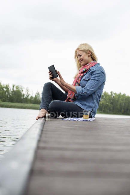 Seitenansicht einer Frau, die auf einem Steg am See sitzt und ein digitales Tablet benutzt. — Stockfoto