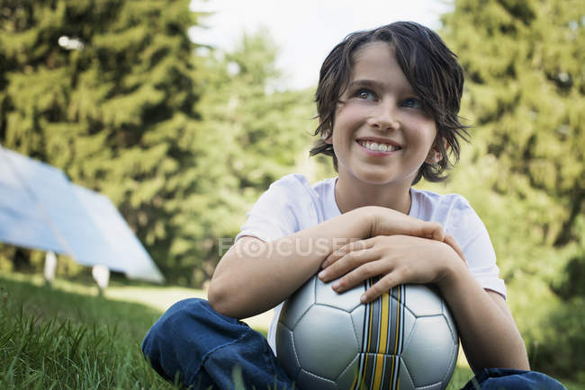Niño sosteniendo el fútbol mientras está sentado en la hierba verde con paneles solares detrás . - foto de stock