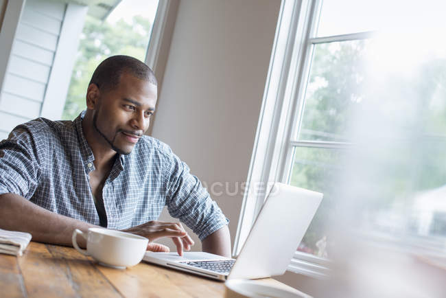 Чоловік використовує ноутбук, сидячи з чашкою кави в кафе . — стокове фото