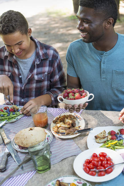 Young man holding bowl of fresh strawberries at picnic table with family in woods. — Stock Photo
