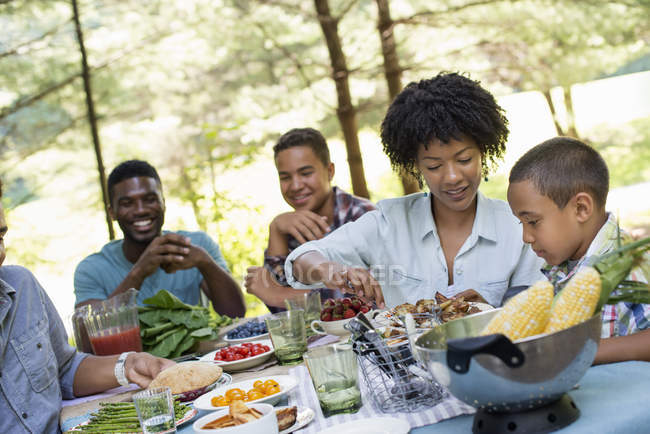 Adultes et enfants assis autour d'une table de pique-nique avec de la nourriture à l'extérieur . — Photo de stock