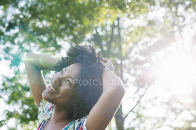 Junge Frau in geblümtem Kleid, die Hände auf dem Kopf, lächelnd und im Wald aufblickend. — Stockfoto