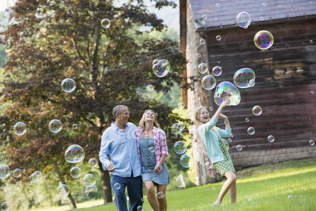 Pais assistindo filha pegando bolhas de sabão e rindo no campo . — Fotografia de Stock