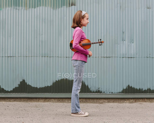 Pre-adolescent girl holding violin on street against corrugated metal wall — Stock Photo