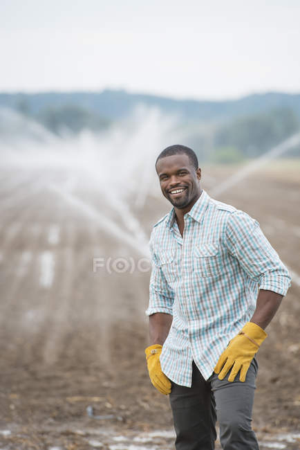 Giovane agricoltore di sesso maschile in abiti da lavoro su campo biologico con irrigazione irrigatori ad acqua . — Foto stock