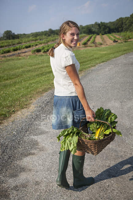 Jeune femme en bottes portant panier de produits frais à la ferme . — Photo de stock