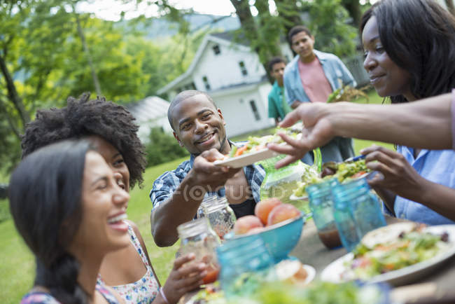Jóvenes amigos compartiendo platos con comida en la mesa de picnic en el jardín rural
. - foto de stock