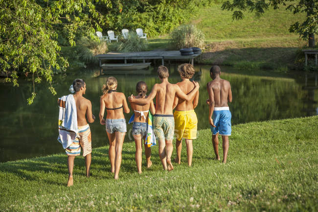 Medium group of teenage boys and girls walking across green field to lake. — Stock Photo