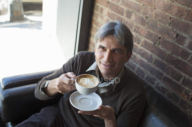 Uomo seduto in caffetteria e con in mano una tazza di cappuccino schiumoso . — Foto stock