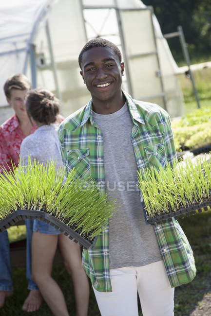 Junger Mann hält Tabletts mit grünen Sämlingen in der Hand, während sich ein Paar vor dem Gewächshaus unterhält. — Stockfoto
