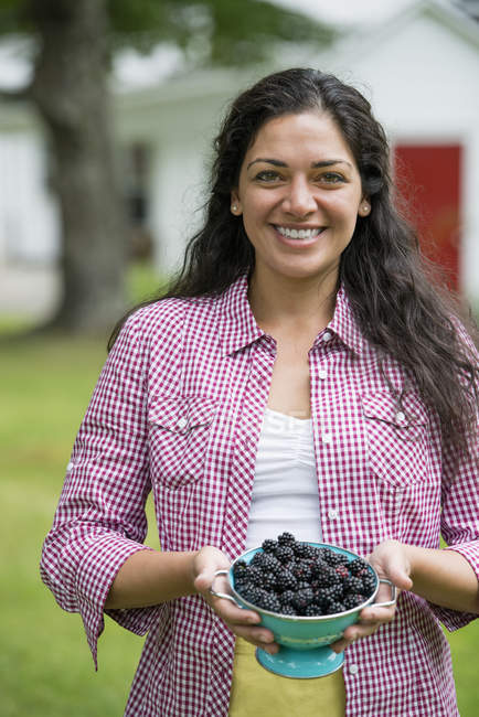 Hispanische Frau mit lockigem Haar hält Schüssel mit frisch gepflückten Brombeeren im Landgarten. — Stockfoto
