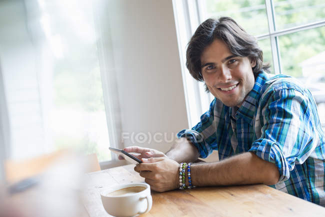 Junger Mann hält digitales Tablet in der Hand und blickt in die Kamera, während er mit einer Tasse Kaffee im Café sitzt. — Stockfoto
