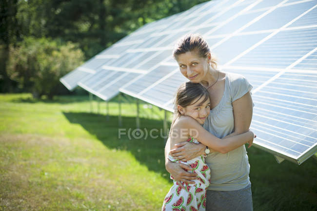 Pre-adolescent girl with mother hugging beside solar panels at farm. — Stock Photo