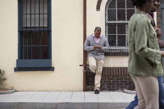Pareja caminando en la acera con el hombre apoyado contra la pared y comprobando el teléfono . - foto de stock
