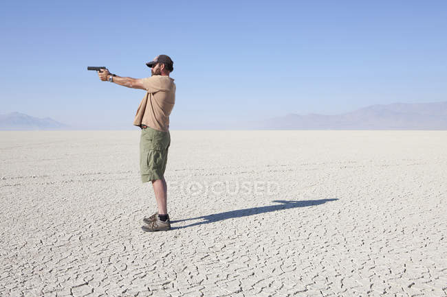Side view of man aiming handgun while standing in vast of barren desert. — Stock Photo