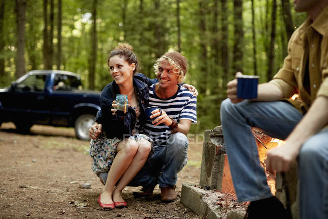 Young friends sitting around campfire at dusk with truck in woods. — Stock Photo