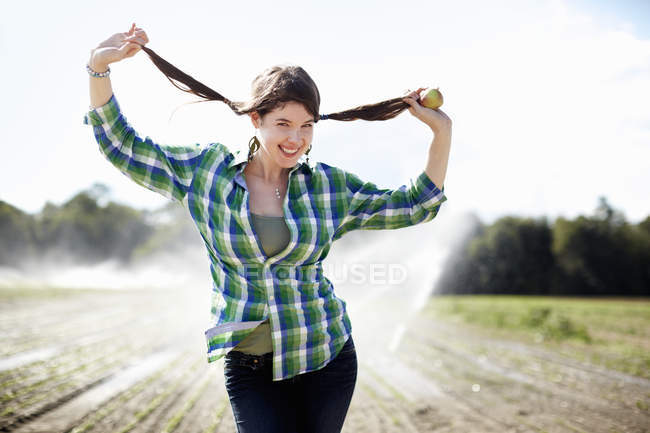 Молода жінка в зеленій перевіреній сорочці тримає коси, стоячи в полі з спринклерами . — стокове фото