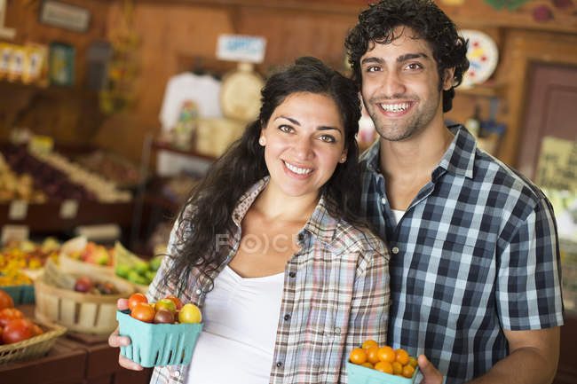 Joven hombre y mujer posando junto con contenedores de tomates maduros en la tienda de agricultores orgánicos . - foto de stock