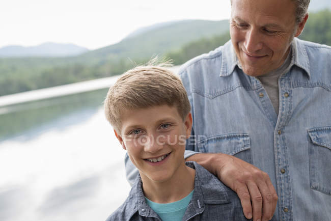 Padre apoyado en el hombro hijo al aire libre en la orilla del lago . - foto de stock