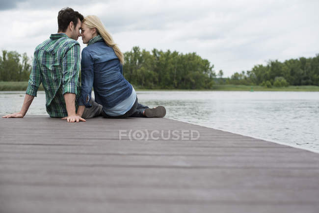 Man and woman sitting head to head on jetty by lake. — Stock Photo