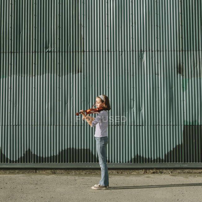 Подростковая девушка играет на скрипке на улице против гофрированной металлической стены — стоковое фото